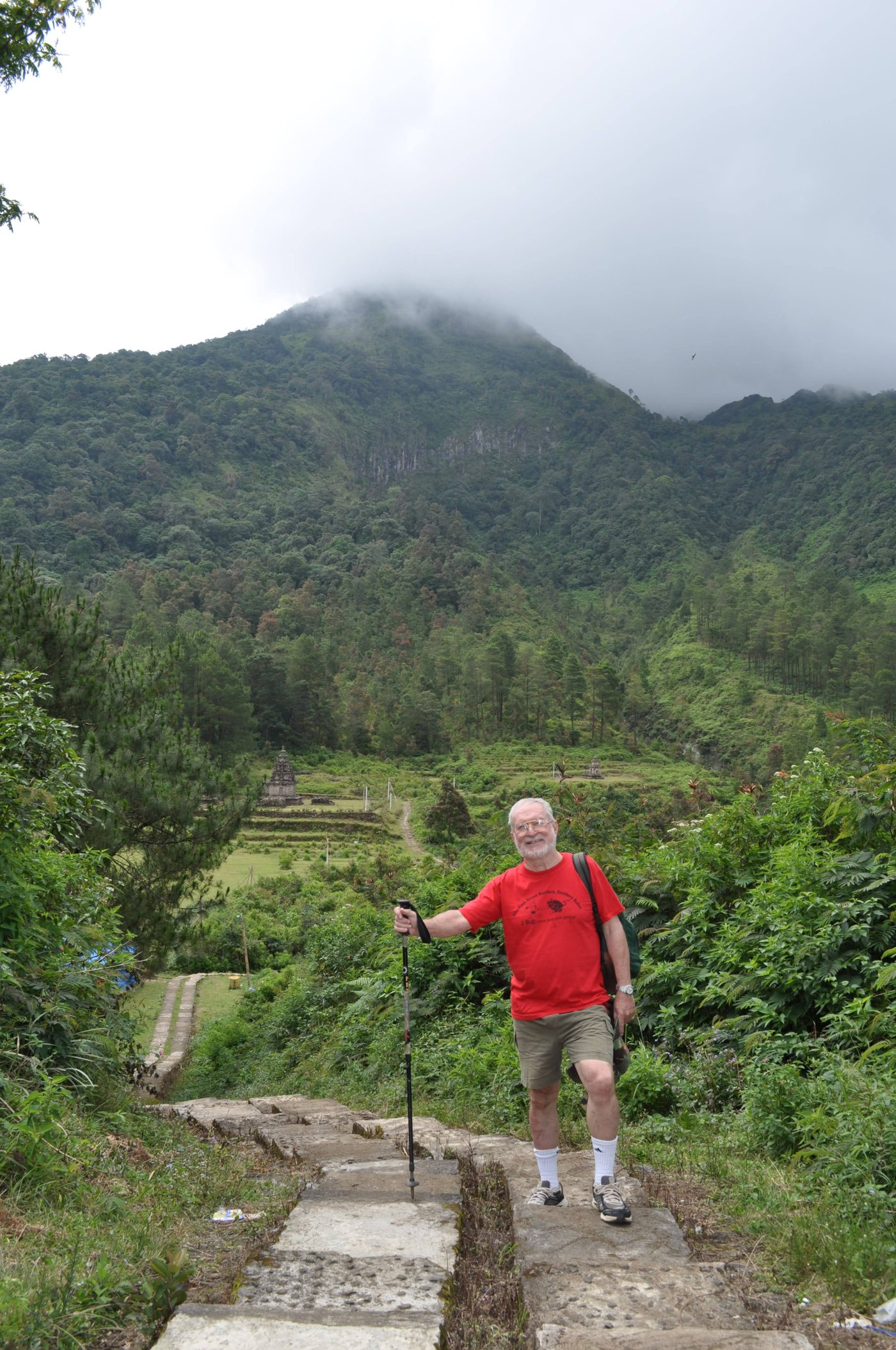 John Wesley Duewel standing on a stone trail high in the green hills, walking pole in hand, with a forested mountain behind him