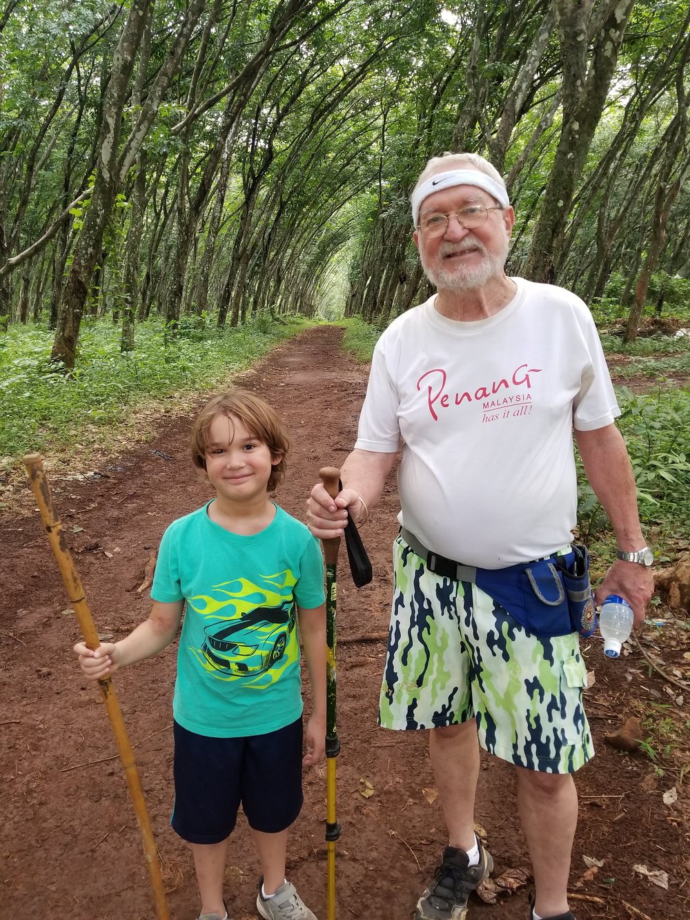 John walking a red earth trail through the trees with one of his grandchildren, both carrying walking sticks