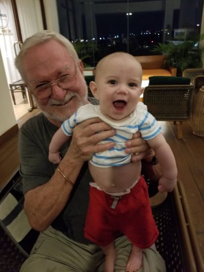 John holding a baby grandchild up to the camera, both delighted
