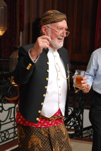 John in Javanese dress, laughing, a glass of beer in his hand