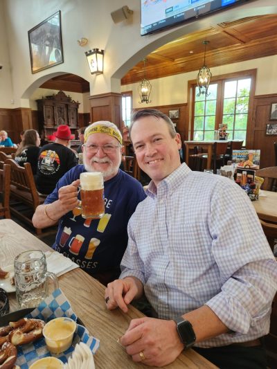 John raising a tall glass of beer with a friend, both mid-laugh