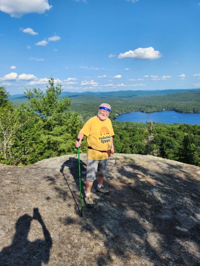 John standing on a bare rock high above a forested lake, walking pole in hand